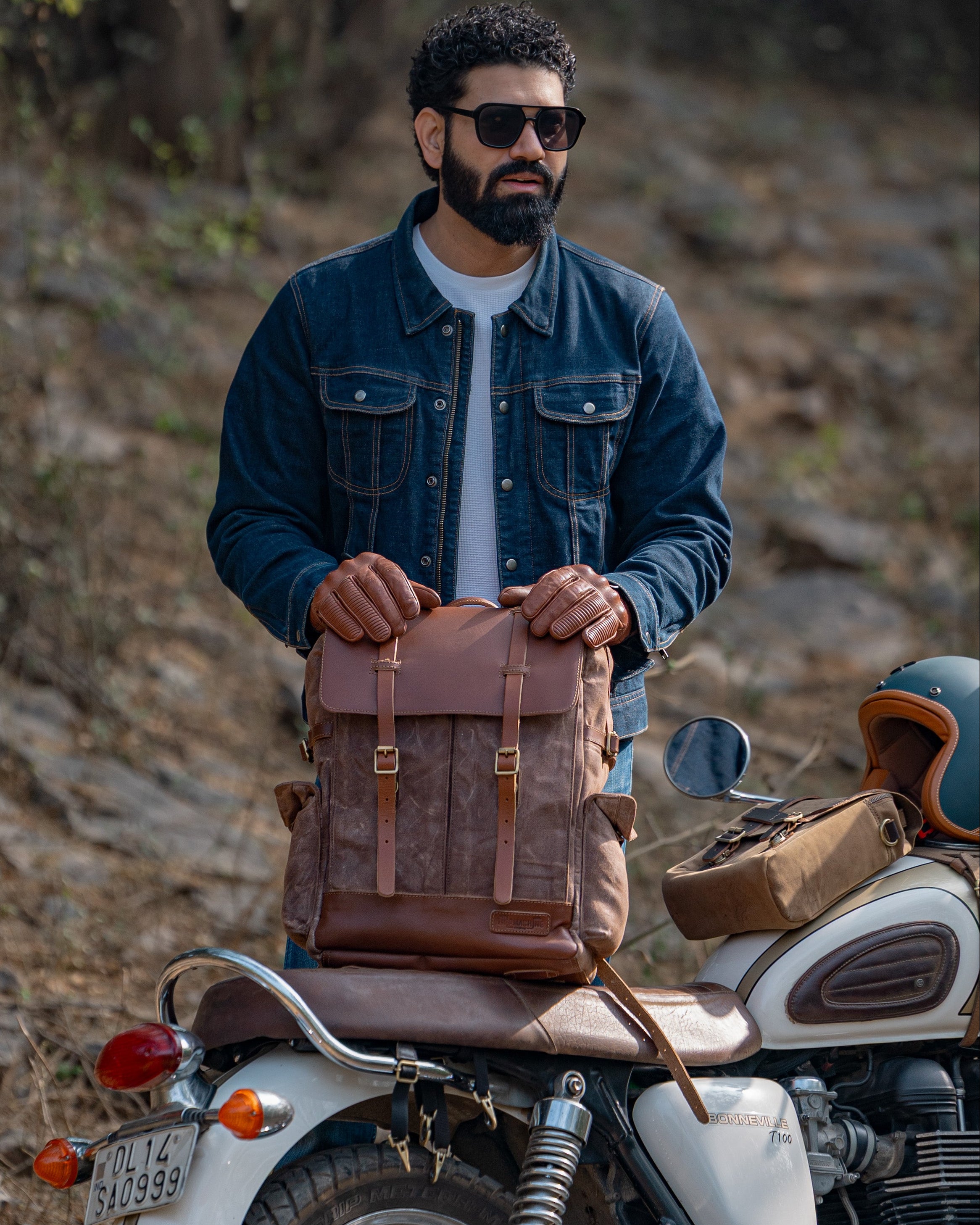Man holding a brown leather bag next to a motorcycle in a forest setting