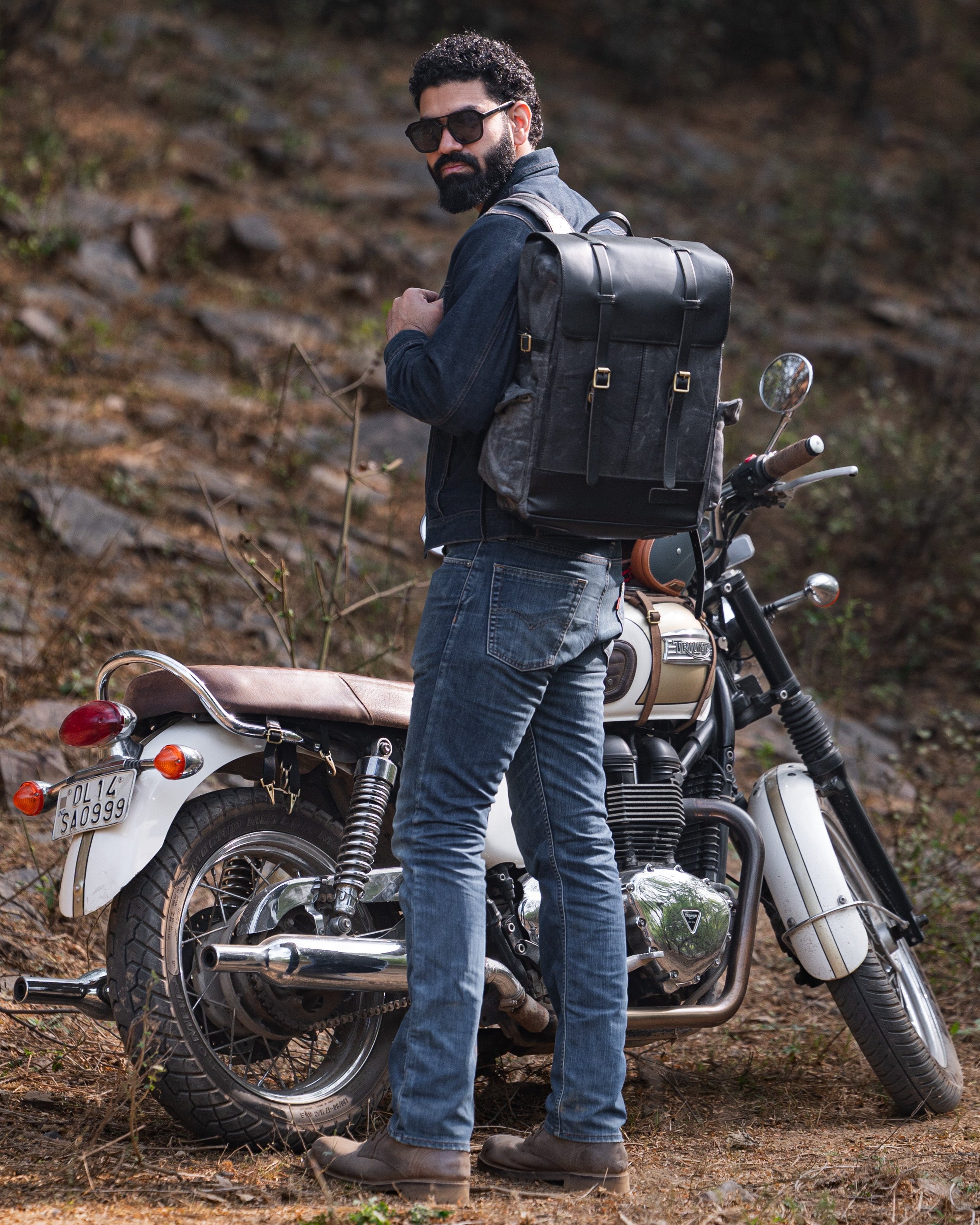 Man with a beard wearing sunglasses and a black leather backpack standing next to a motorcycle in a natural setting.
