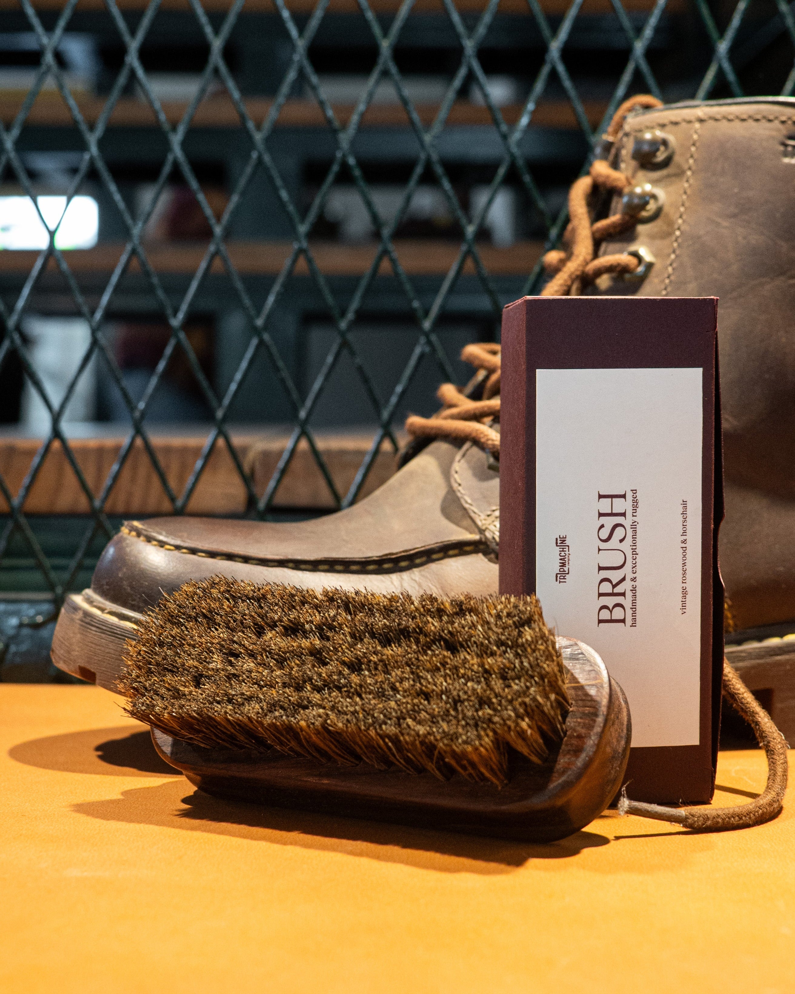 Brown leather boot with a brush and care card on an orange surface, against a metal grid background.