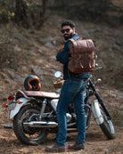 Man with a brown leather backpack standing next to a motorcycle in a forest setting