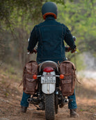 Person riding a motorcycle with saddlebags on a dirt road in a forest.