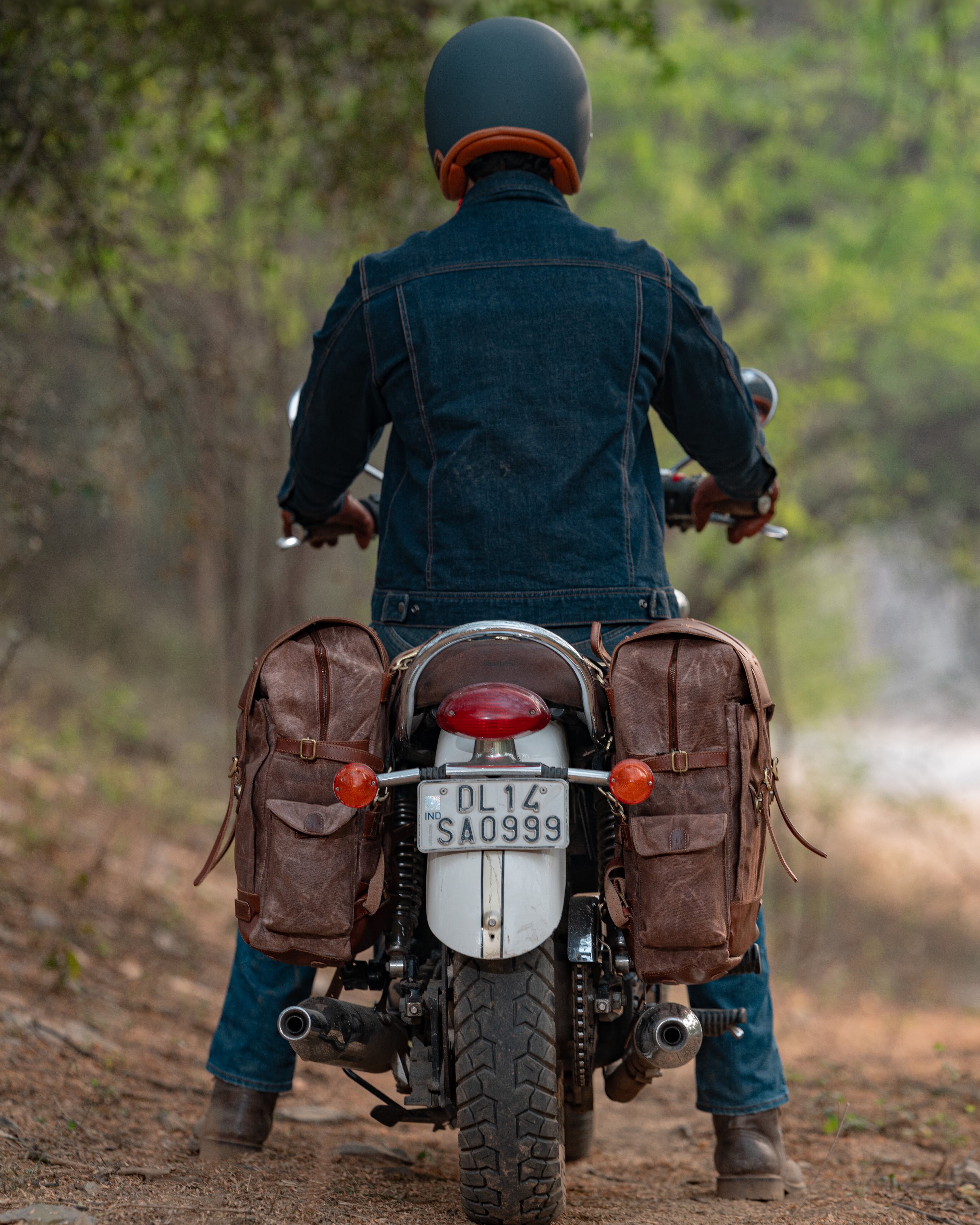 Person riding a motorcycle with saddlebags on a dirt road in a forest.