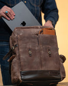 Person holding a laptop next to a brown leather backpack on a wooden surface.