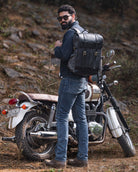 Man with a beard wearing sunglasses and a black leather backpack standing next to a motorcycle in a natural setting.