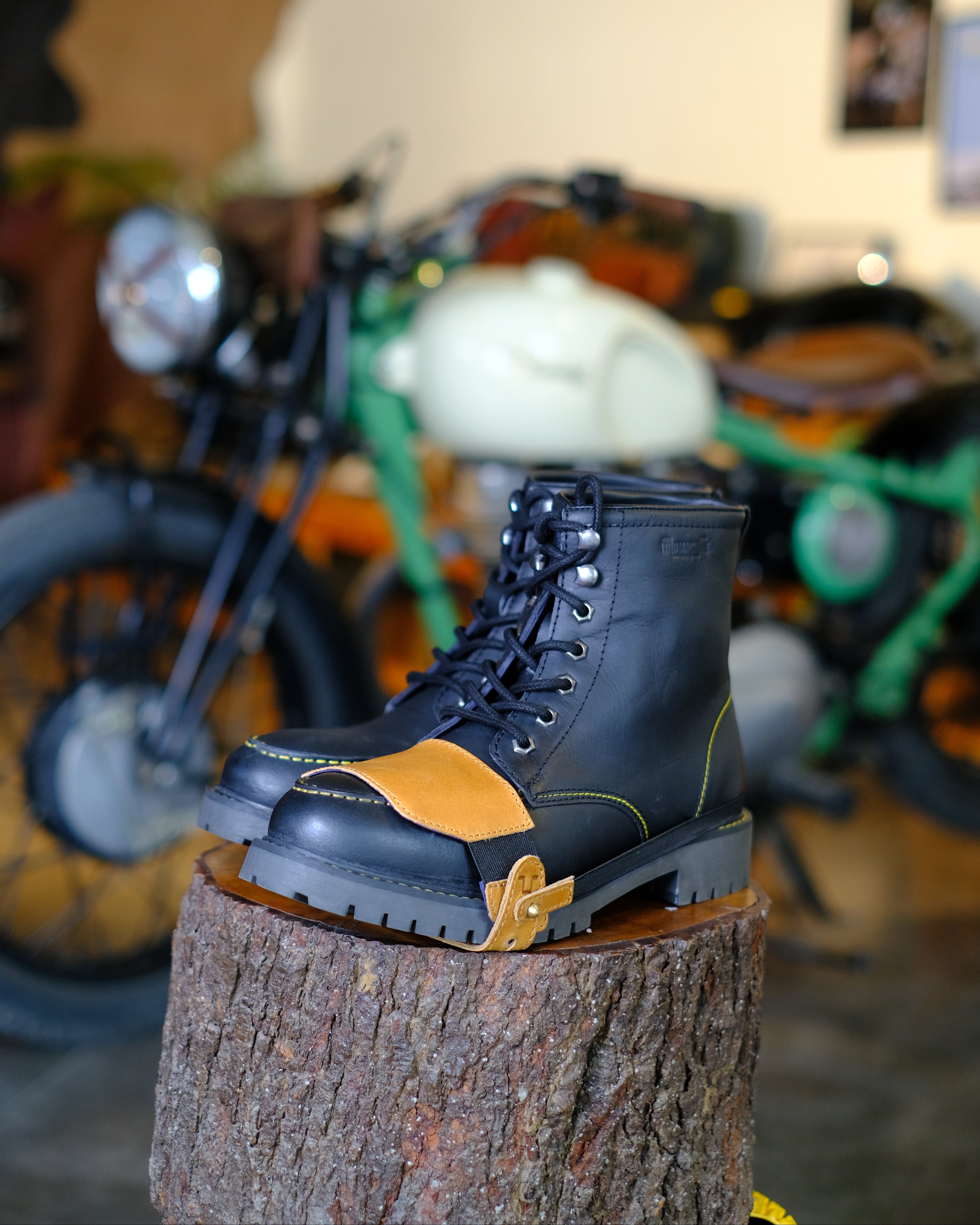 Black boot with tan shoe protector on a wooden stump with a motorcycle in the background