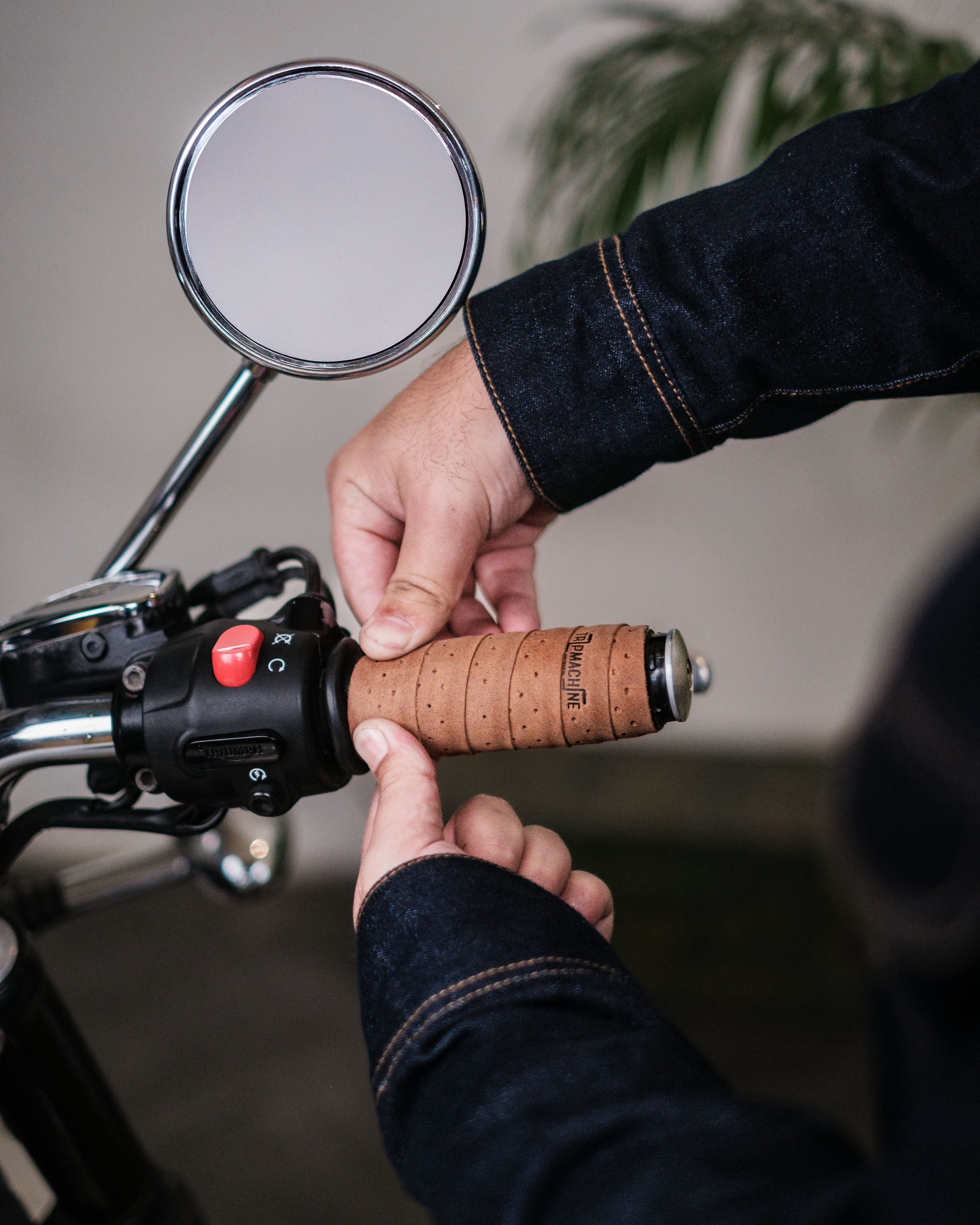 A person's hand is shown wrapping a brown leather grip wrap onto the handlebars of a motorcycle, with a focus on the grip and handlebar area.